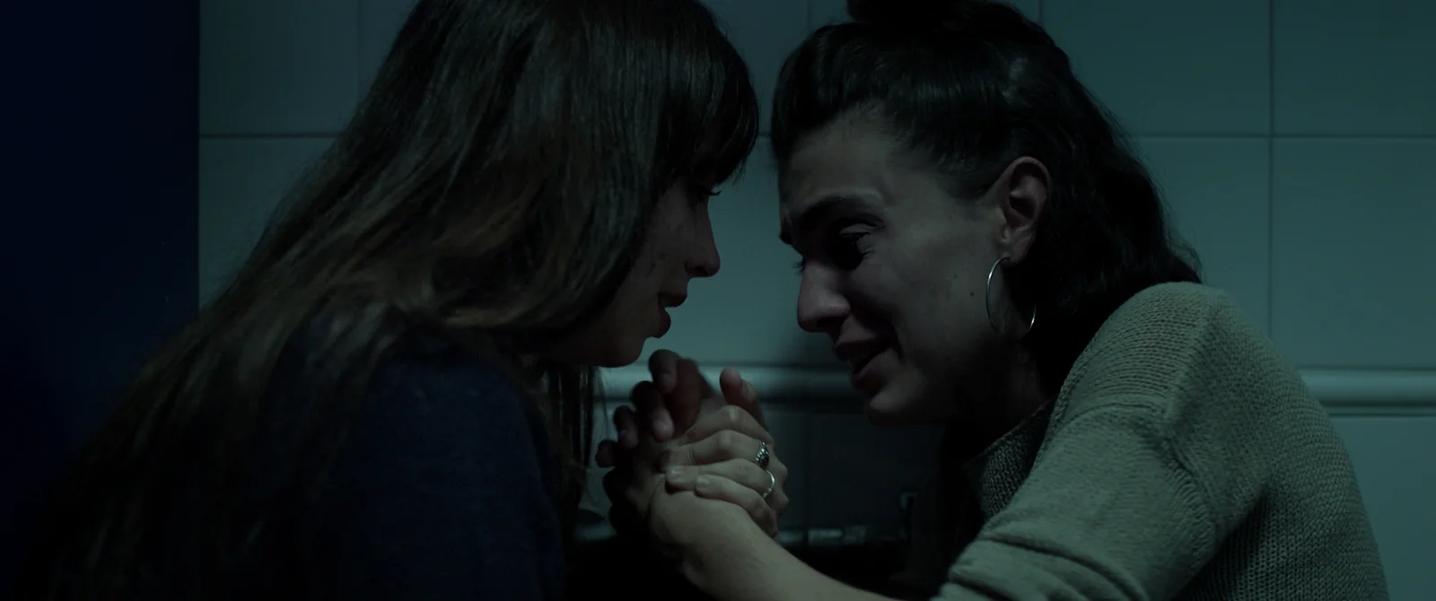 Dos mujeres se dan la mano en una estancia alicatada y oscura; una de ellas, con aro en la oreja, llora.
