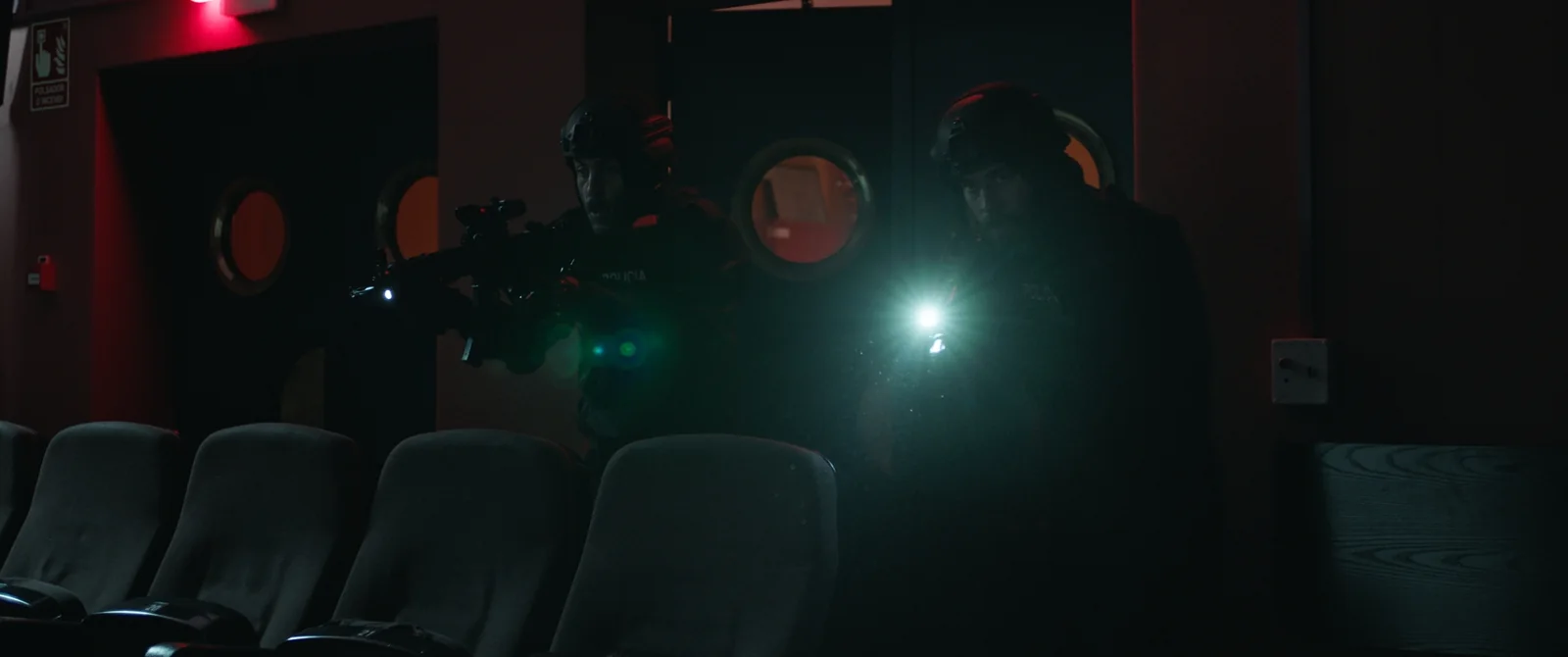 Agentes armados con fusiles y linternas avanzan entre butacas vacías de una sala de cine, en penumbra roja.