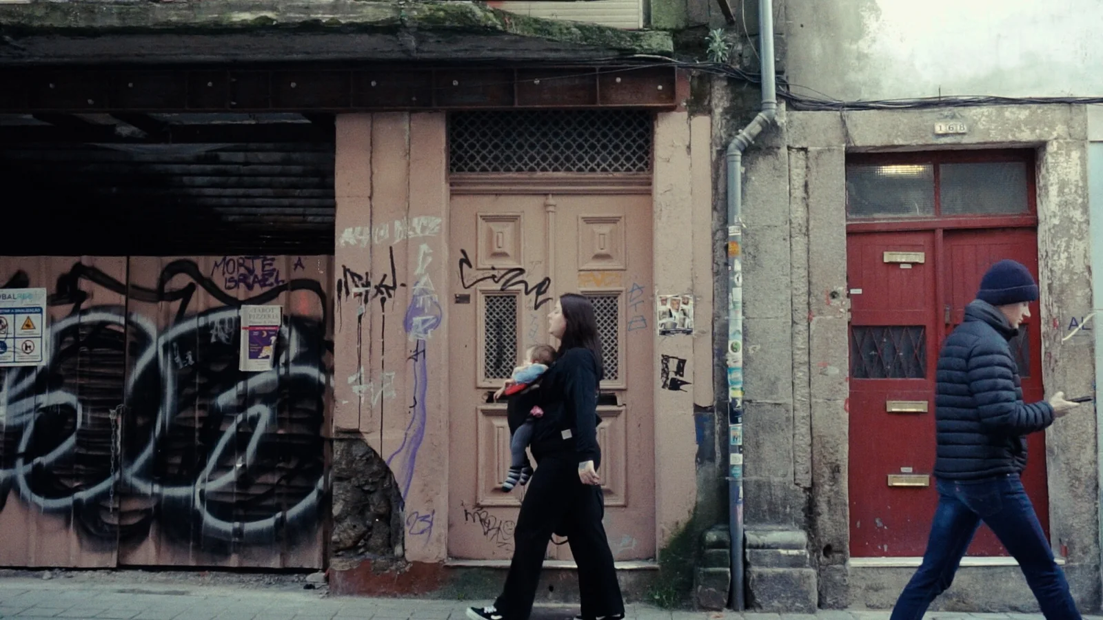 Calle de fachadas desgastadas con grafitis: una mujer de negro lleva a un bebé y un hombre de gorro azul camina.