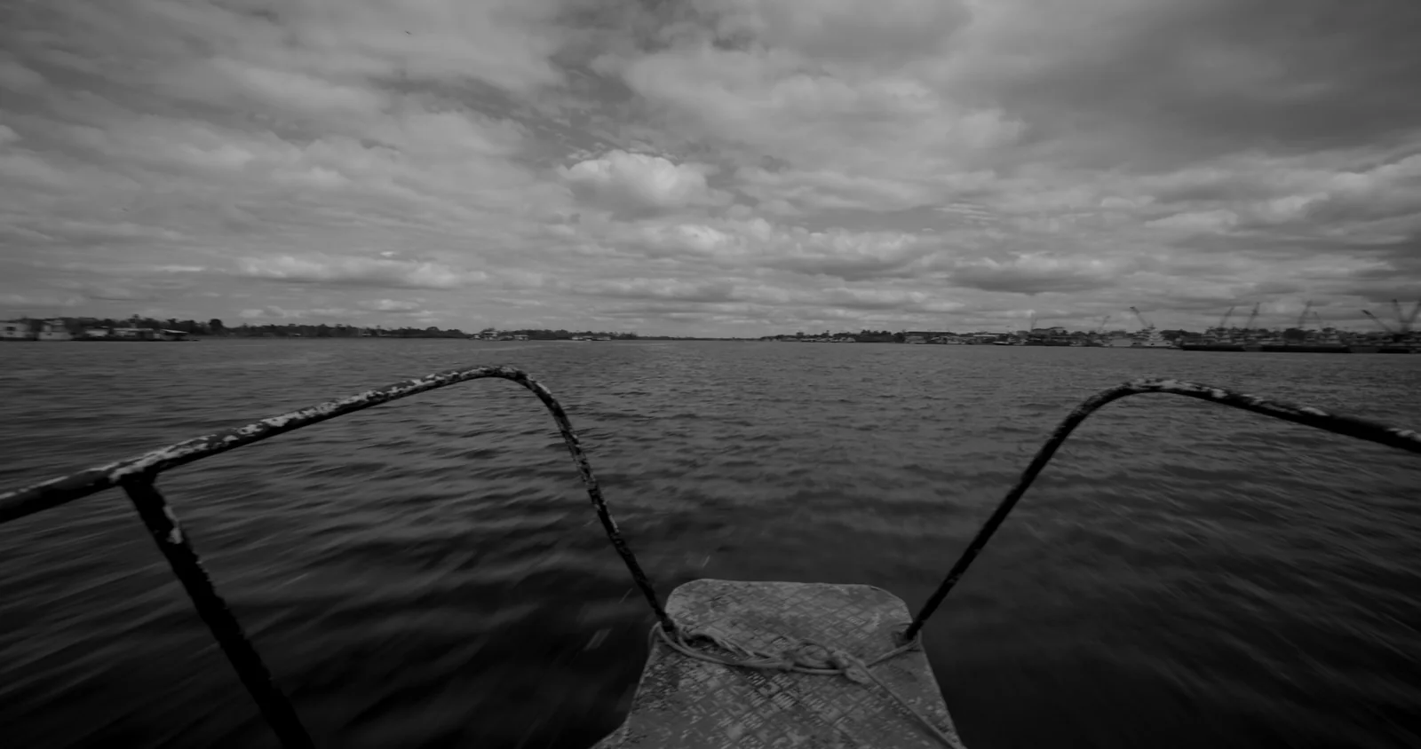 Blanco y negro de la proa de una barca con barandillas metálicas, avanzando por un río bajo un cielo nublado.