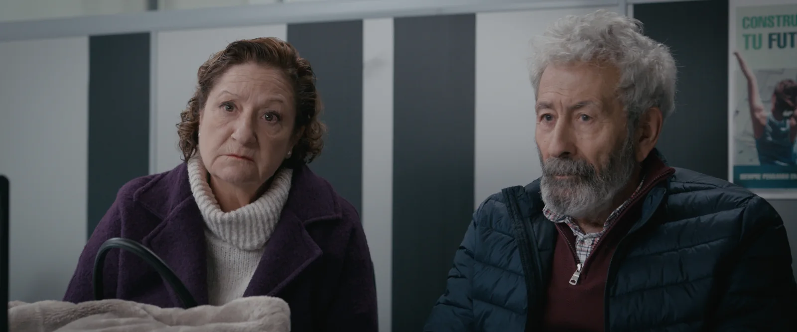 Matrimonio de edad madura sentado en una oficina: ella con abrigo morado y él con chaqueta acolchada azul.