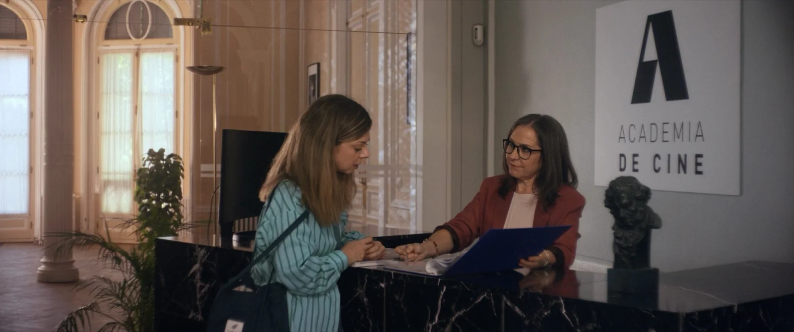 Mujer de gafas y chaqueta roja tras un mostrador de mármol negro, junto a un cartel «Academia de cine».
