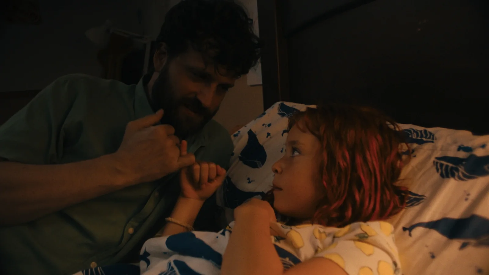 Hombre barbudo de camiseta verde y niña pelirroja se dan el meñique tumbados en una cama, de noche.