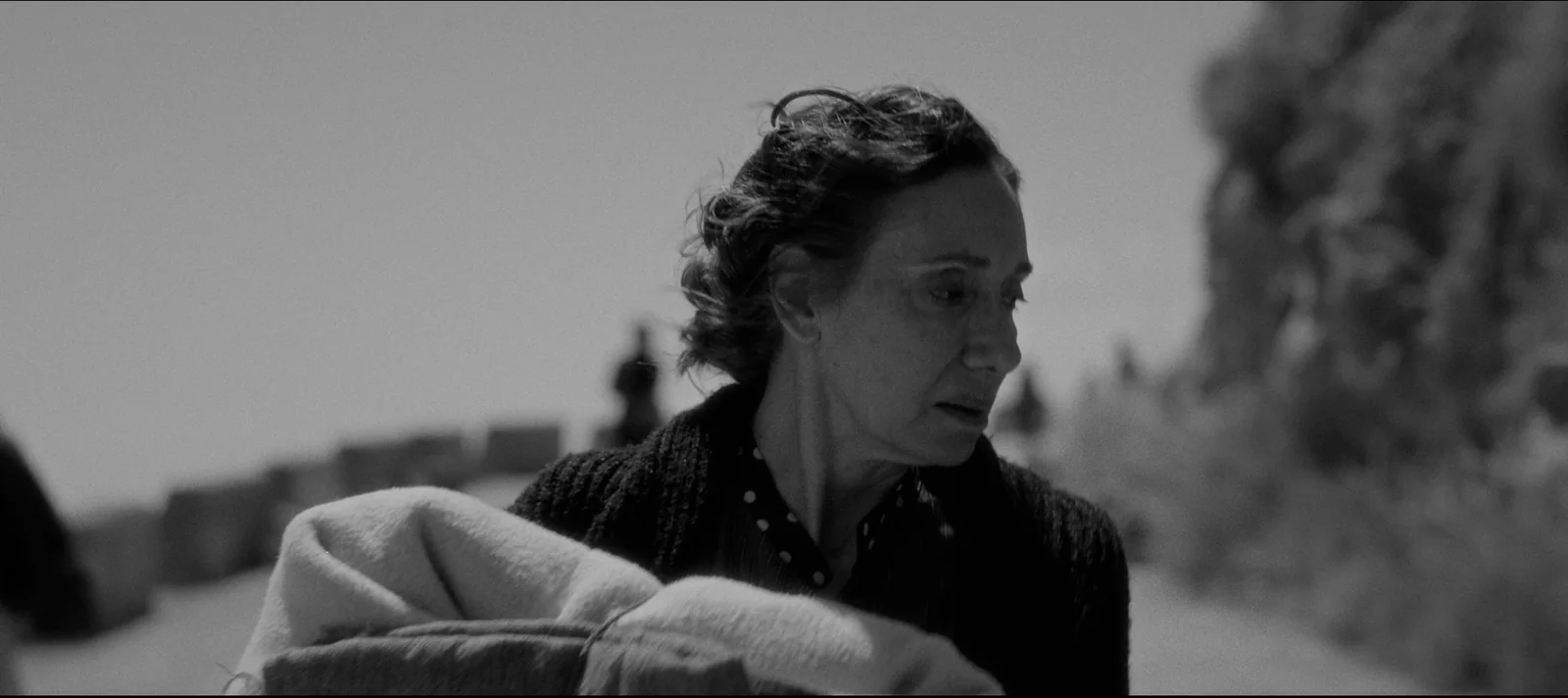Plano en blanco y negro de una mujer mayor de pelo ondulado y abrigo oscuro, mirando hacia un lado junto al mar.