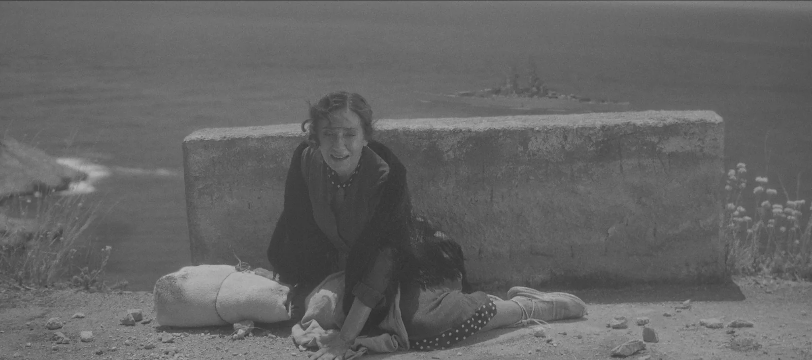 Mujer llorando en blanco y negro, sentada junto a un muro frente al mar, con una niña tendida en el suelo junto a ella.