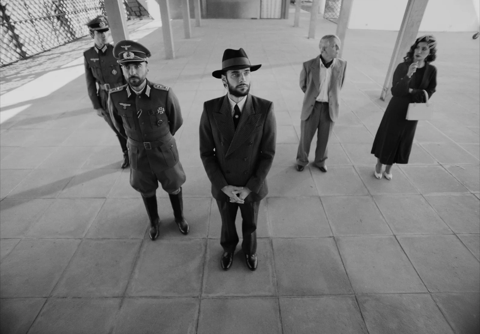 Blanco y negro: un grupo de hombres de uniforme y traje en una terraza; en el centro, uno con sombrero y traje cruzado.