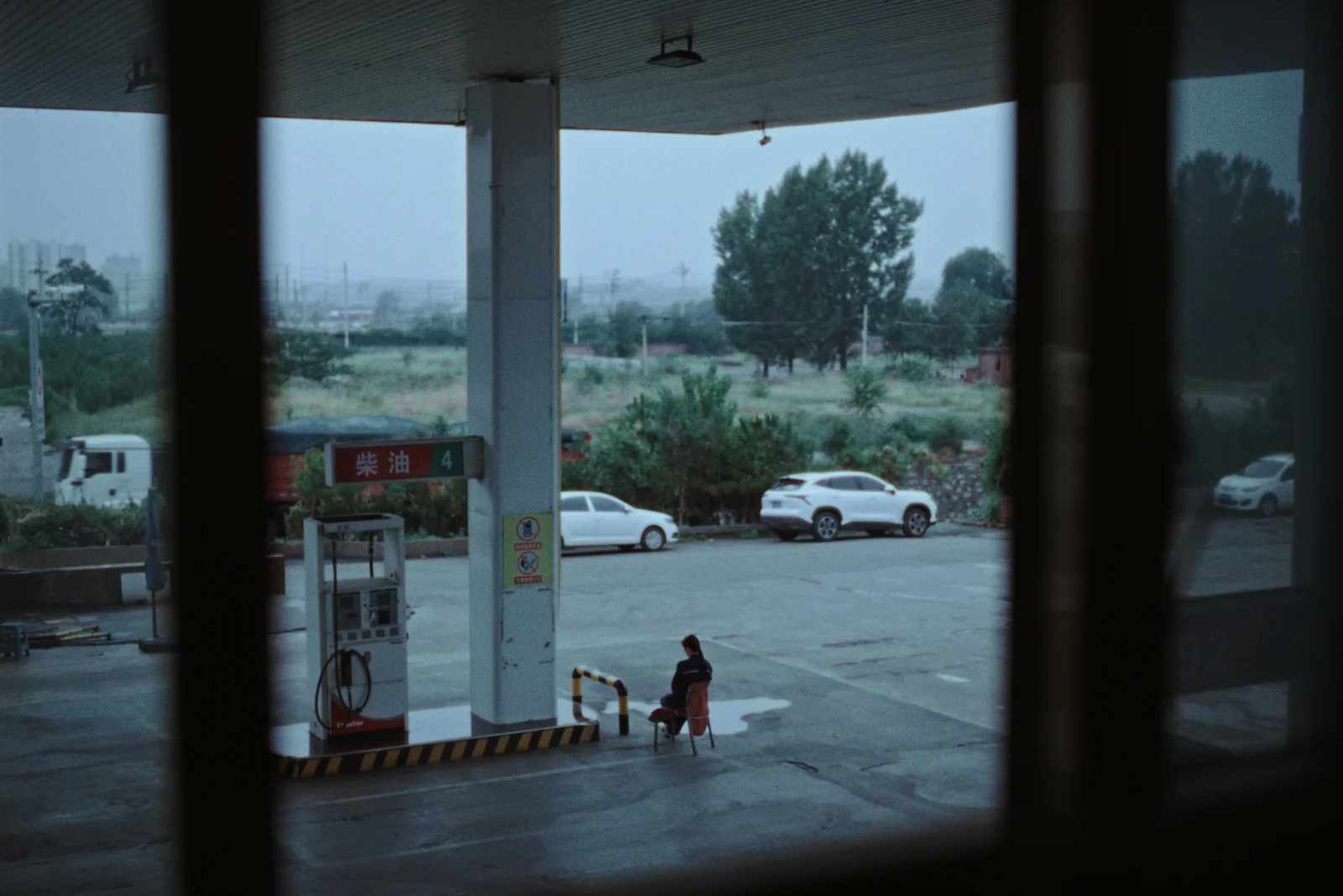 Gasolinera vacía bajo la lluvia, con una persona sentada junto a un pilar y un cartel en chino de diésel.