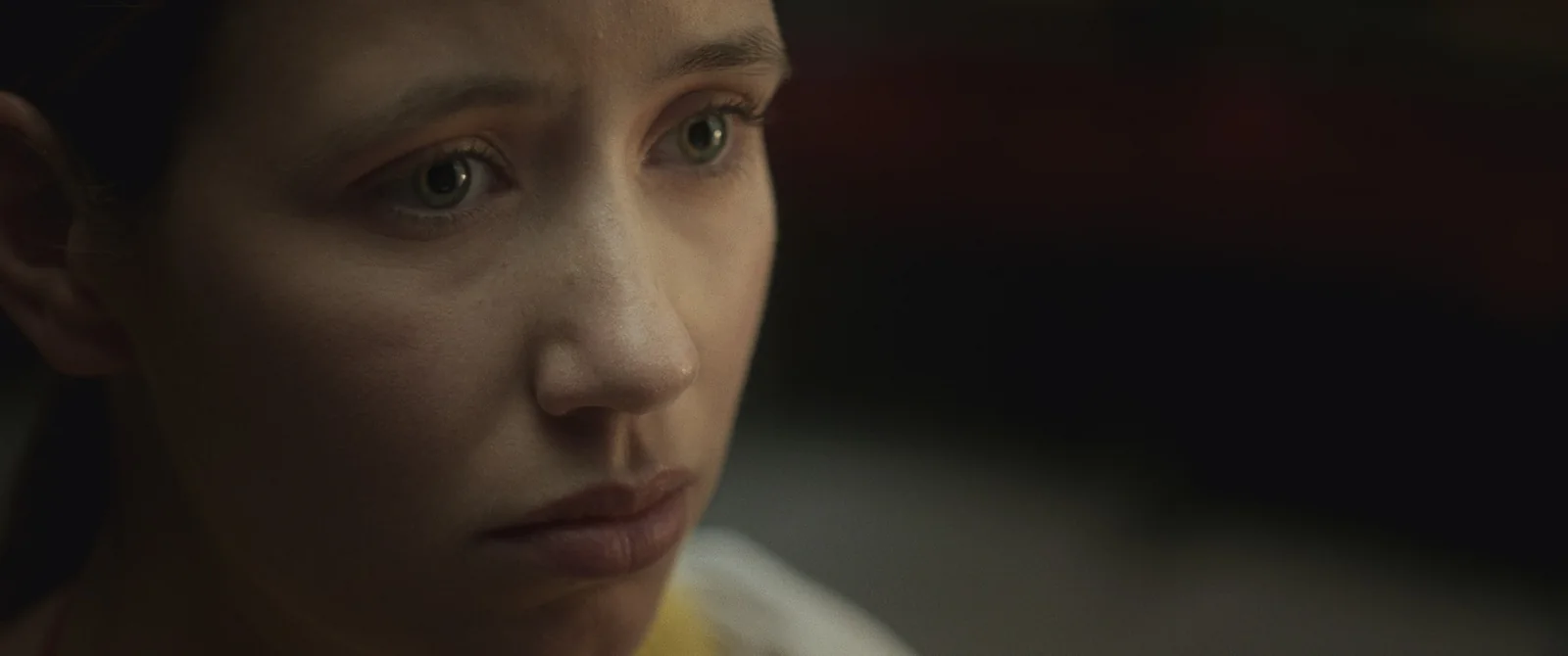 Primer plano de una joven de piel clara y ojos verdosos, con gesto triste, en un ambiente oscuro.