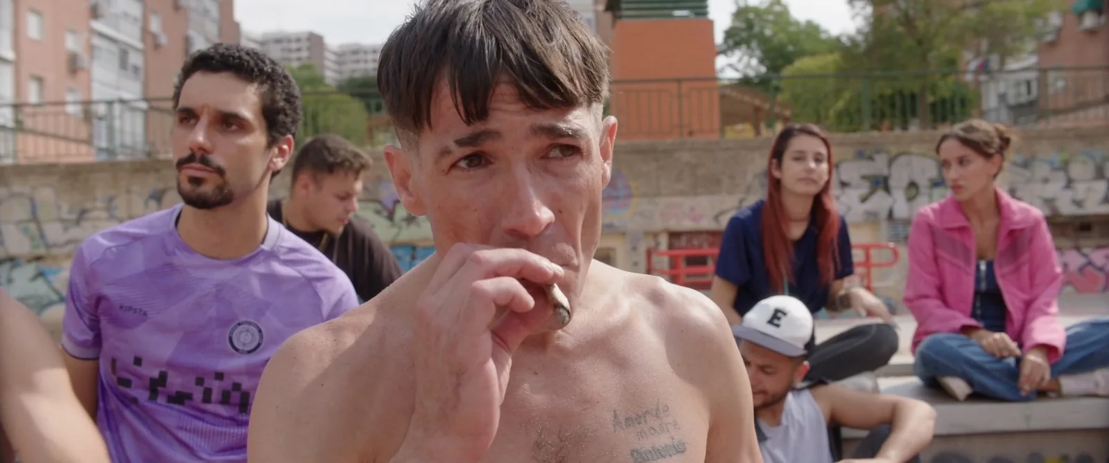 Hombre sin camiseta, con un tatuaje en el pecho, llevándose un cigarrillo a la boca junto a jóvenes sentados.
