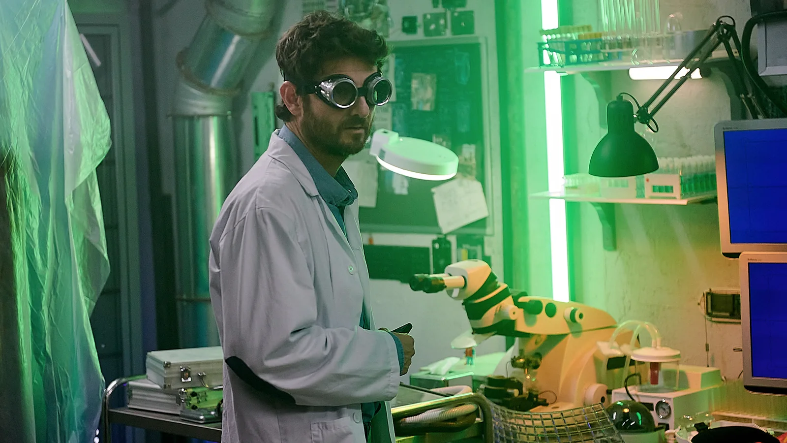 Hombre con bata blanca y gafas de protección en un laboratorio iluminado en verde, junto a un brazo robótico.