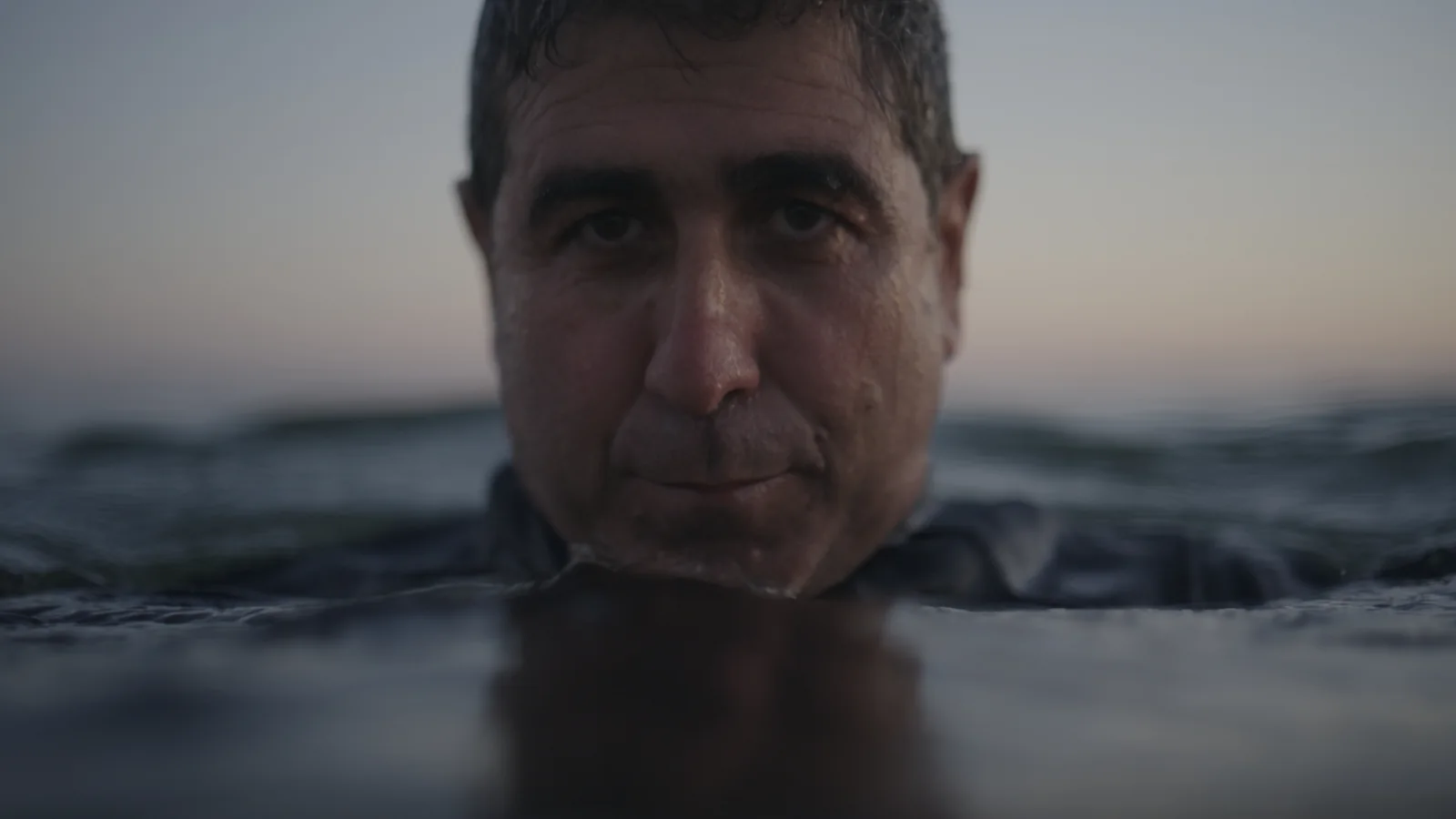 Hombre de pelo corto que emerge del agua hasta la nariz, mirando fijo a cámara con el atardecer al fondo.