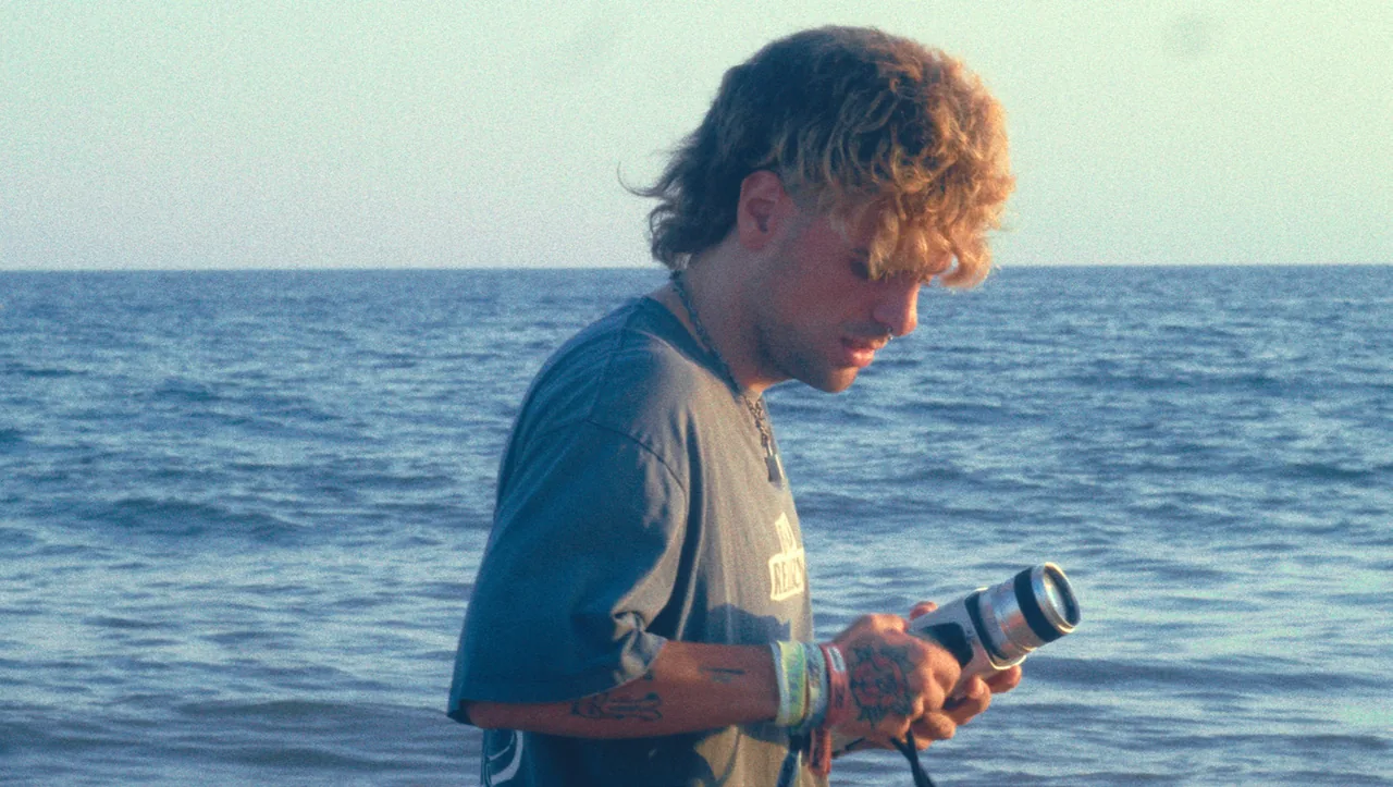 Fotograma de «Herencia»: un joven sostiene una cámara de cine antigua a la orilla del mar.