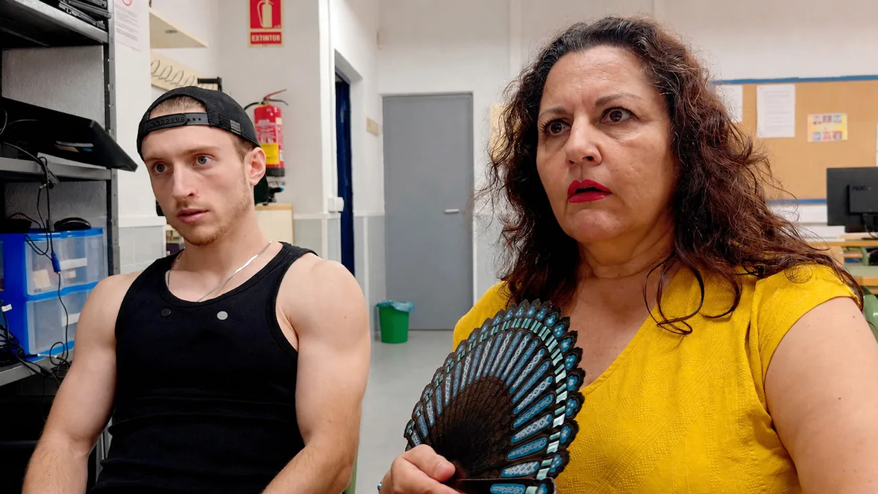 Fotograma de «Una tutoría entretenida»: un joven con gorra y camiseta de tirantes y una mujer con abanico, sentados en un despacho escolar.
