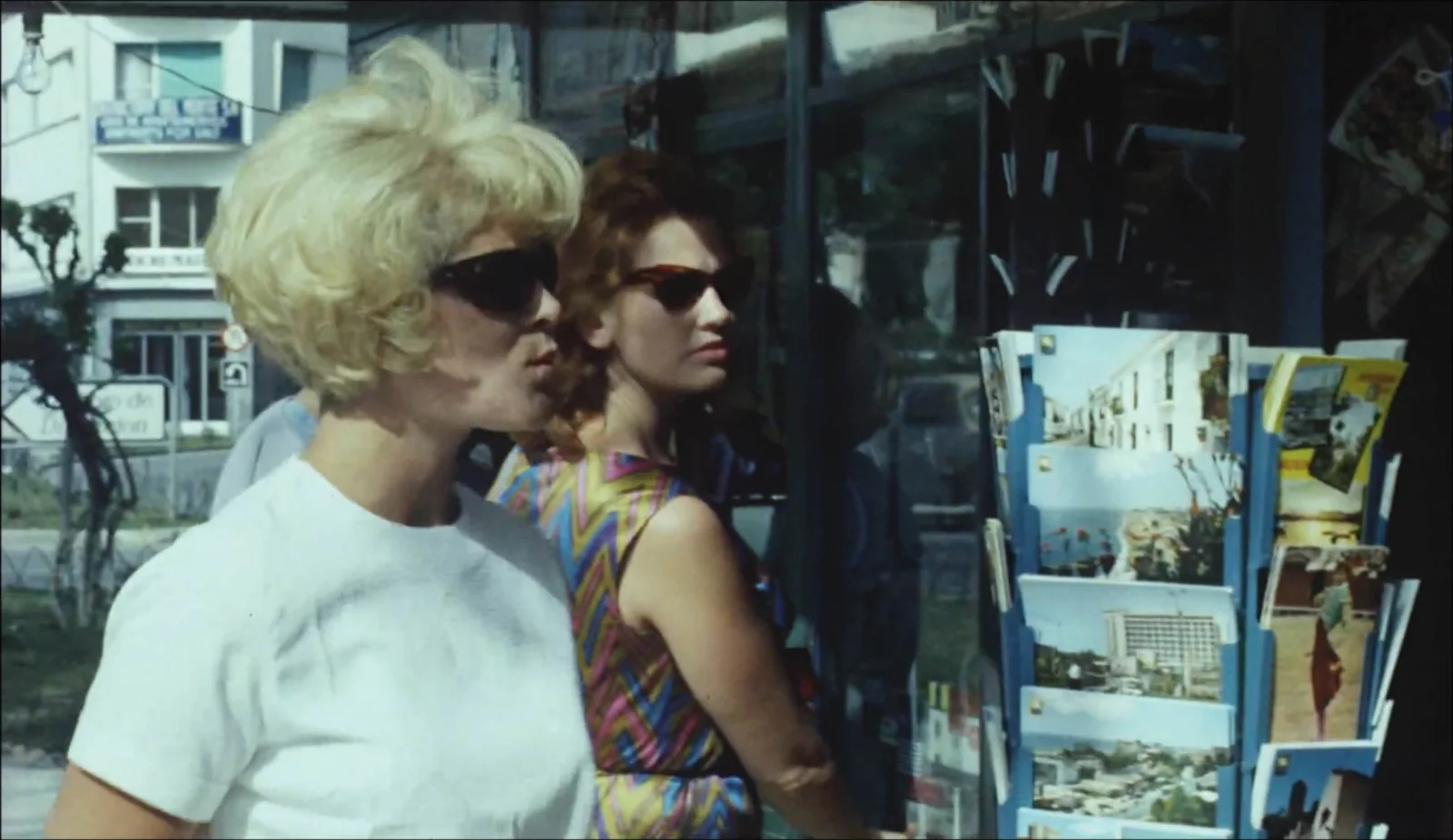 Fotograma de «Back in Time!»: dos mujeres rubias con gafas de sol, en los años sesenta, junto a un expositor de postales.