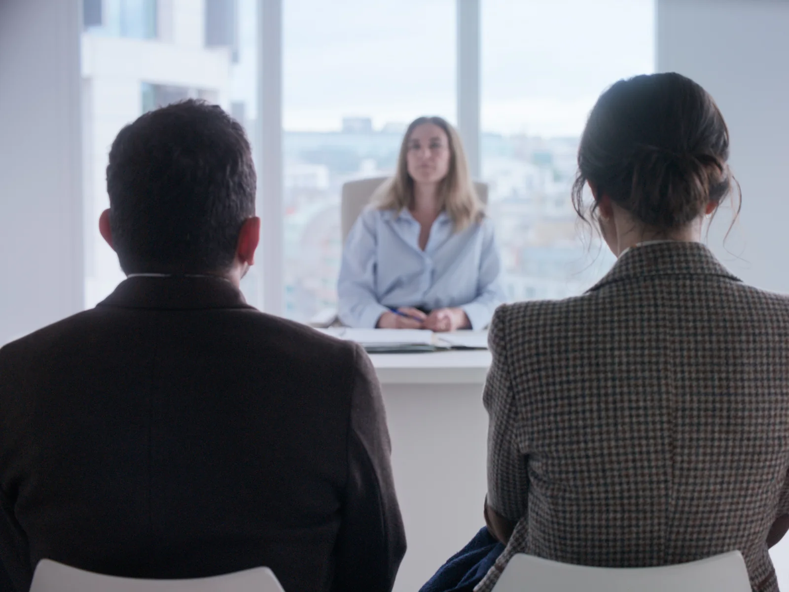 Fotograma de «On vous croit»: una mujer sentada tras una mesa, en una sala luminosa con grandes ventanales, frente a dos personas de espaldas.