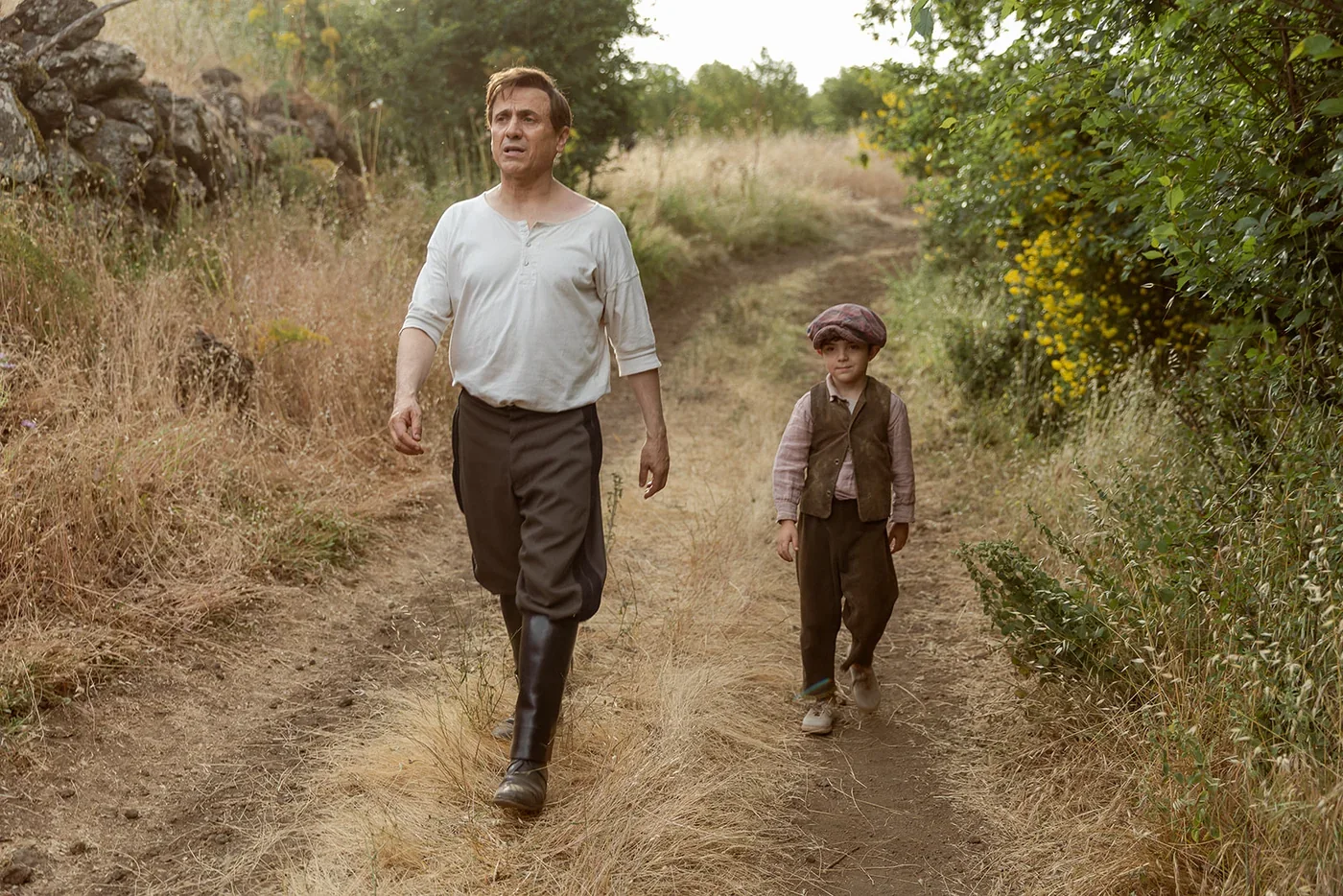 Fotograma de «Arriba tutto»: un hombre con camisa blanca y botas camina por un sendero de campo junto a un niño con gorra.