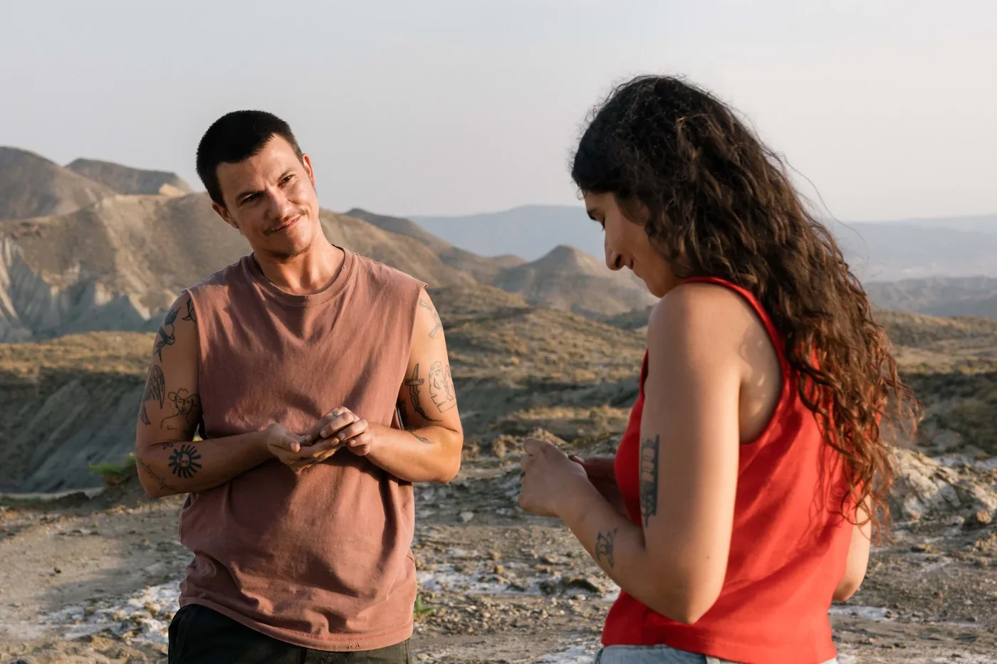 Fotograma de «Iván & Hadoum»: un joven con camiseta sin mangas y una mujer de pelo rizado conversan ante un paisaje desértico.