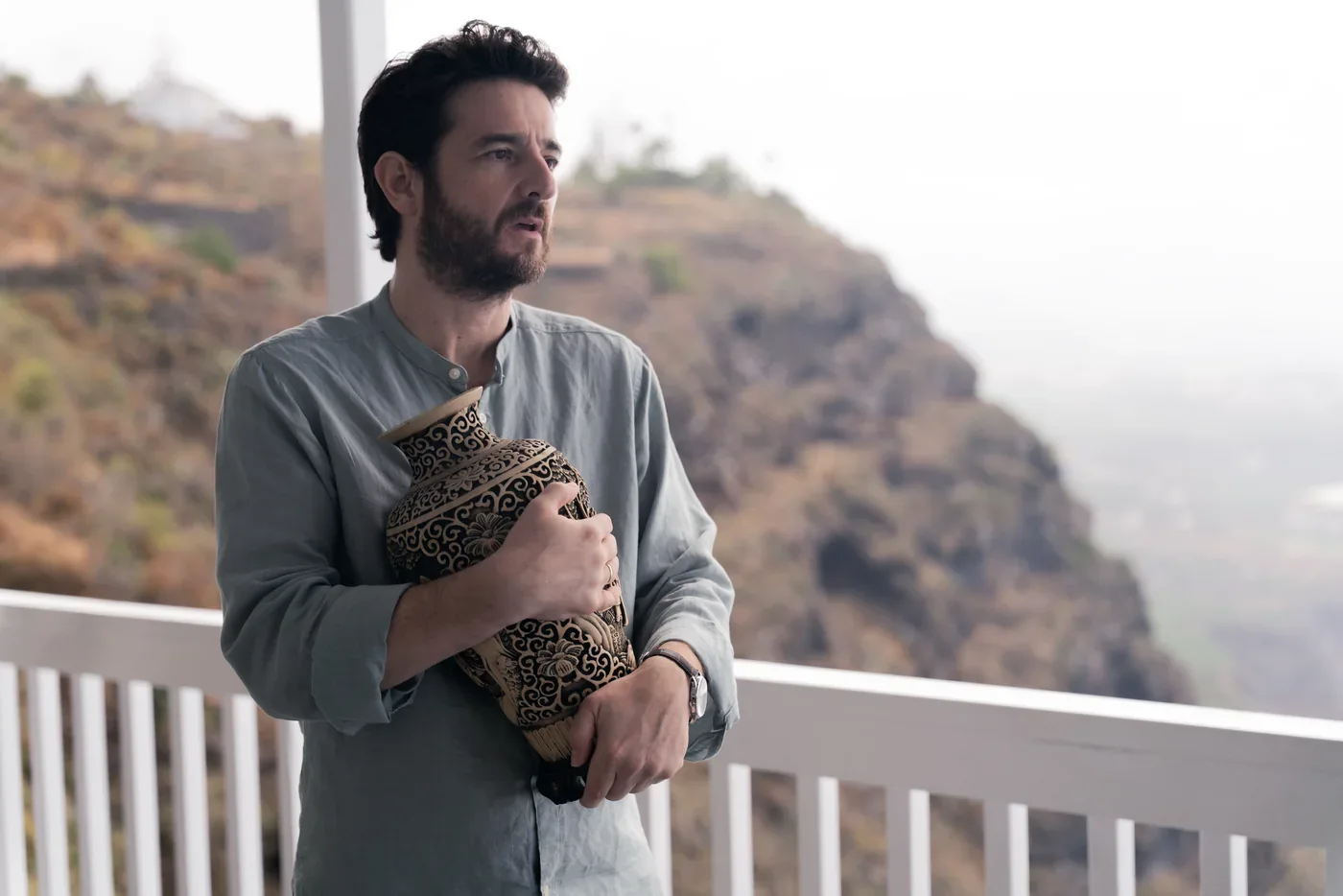 Fotograma de «Ni contigo ni sin mí»: un hombre con barba sostiene un jarrón en una terraza con vistas a la montaña.
